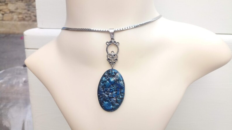 May include: A silver-toned necklace with an oval, blue pendant. The pendant has a textured, mosaic-like appearance. The necklace is displayed on a mannequin bust, showcasing the jewelry's design and details.
