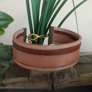 May include: A brown leather bracelet with a gold clasp. The bracelet is made of two layers of leather, with a brown stripe running along the edge of the top layer.