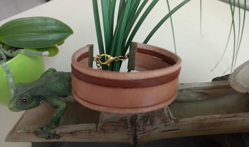 May include: A brown leather bracelet with a gold clasp. The bracelet is made of two layers of leather, with a brown stripe running along the edge of the top layer.