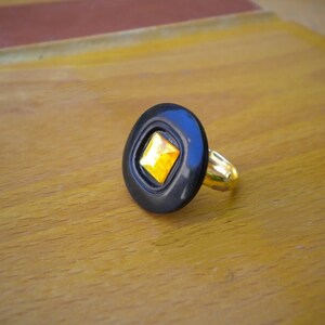 Puede incluir: Un anillo negro con una banda dorada y una gema cuadrada amarilla engastada en un engaste negro de forma ovalada. El anillo se muestra sobre una superficie de madera.