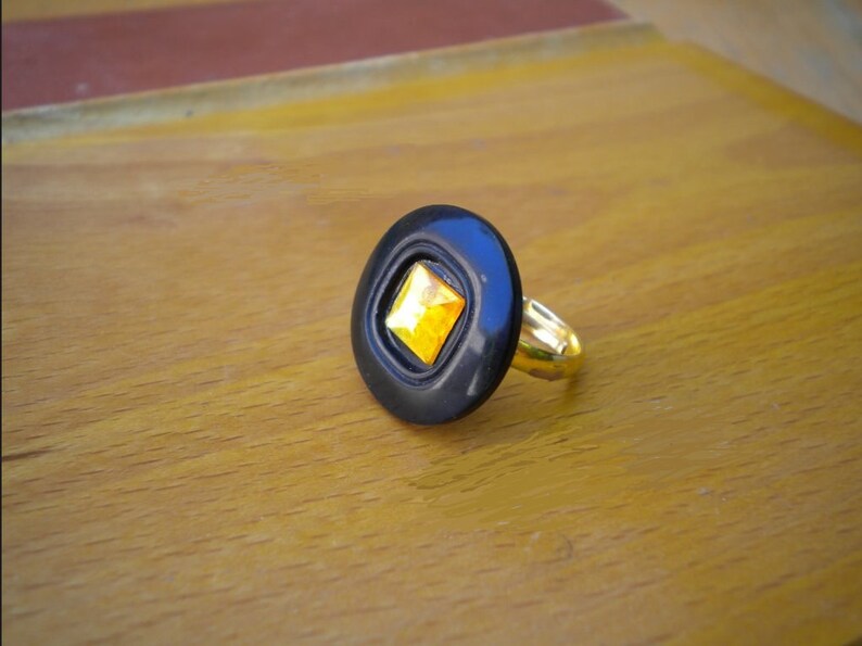 Puede incluir: Un anillo negro con una banda dorada y una gema cuadrada amarilla engastada en un engaste negro de forma ovalada. El anillo se muestra sobre una superficie de madera.