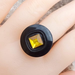 Puede incluir: Un anillo negro con un dise&ntilde;o circular y una gema amarilla cuadrada en el centro. El anillo se encuentra sobre un fondo neutro, resaltando su color y forma. El anillo es una pieza de joyer&iacute;a.