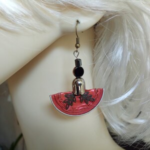 Puede incluir: Pendientes rojos y plateados con cuentas negras y un diseño de hojas. Los pendientes cuelgan de la oreja de una persona.