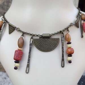 Puede incluir: Un collar de cuentas marrones y rojas con un gran colgante circular de metal. El colgante tiene un diseño texturizado de círculos concéntricos. El collar también presenta varios abalorios y cuentas metálicas más pequeños.