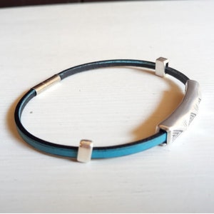 May include: A bracelet with a black and turquoise leather strap and silver-coloured metal accents. The bracelet features a curved, engraved centrepiece and two square end pieces. The clasp is also silver-coloured.