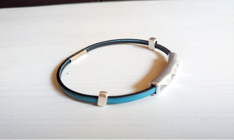 May include: A bracelet with a black and turquoise leather strap and silver-coloured metal accents. The bracelet features a curved, engraved centrepiece and two square end pieces. The clasp is also silver-coloured.