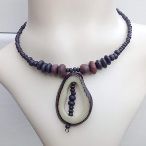 Peut inclure: Un collier de perles brun et noir avec un grand pendentif de forme ovale en bois brun clair avec un centre sculpté. Le pendentif contient une rangée de petites perles noires à l'intérieur du centre sculpté.