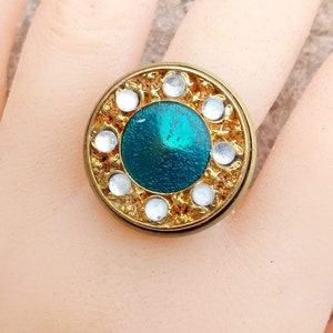 Puede incluir: Anillo dorado con diseño circular. El centro presenta una piedra de color turquesa, rodeada de cristales redondos y transparentes. El borde exterior tiene un patrón texturizado y adornado. Un anillo llamativo de estilo vintage.