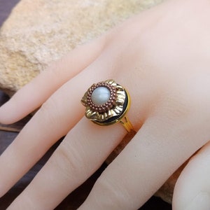 May include: A gold-toned ring featuring a central pearl surrounded by a floral design with red and gold accents. The ring is set on a gold band. The ring is displayed on a hand.