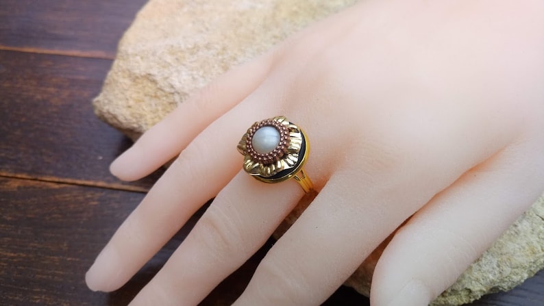 May include: A gold-toned ring featuring a central pearl surrounded by a floral design with red and gold accents. The ring is set on a gold band. The ring is displayed on a hand.