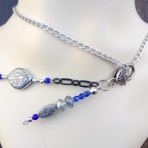 Może przedstawiać: Srebrny naszyjnik z łańcuszkiem i ozdobnymi zawieszkami. Naszyjnik posiada kwadratowy charms, czarne ogniwa łańcucha i niebieskie koraliki. Naszyjnik jest prezentowany na manekinie.