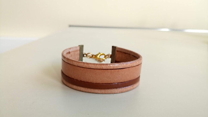 May include: A light brown leather bracelet with a brown leather band and a gold clasp.