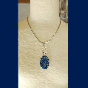 May include: Silver-toned necklace with an oval pendant. The pendant features a blue and silver speckled design. The necklace is displayed on a mannequin. The pendant is approximately 1.5 inches long.