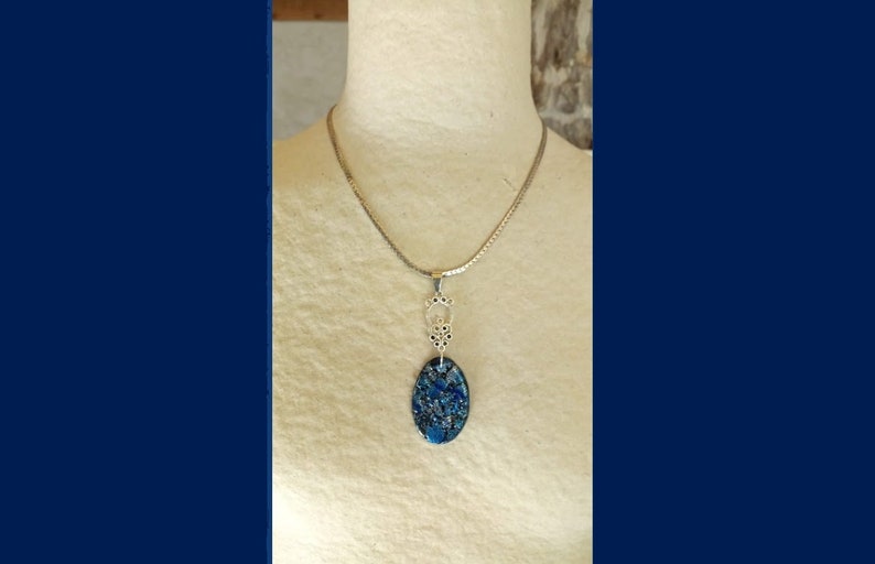 May include: Silver-toned necklace with an oval pendant. The pendant features a blue and silver speckled design. The necklace is displayed on a mannequin. The pendant is approximately 1.5 inches long.