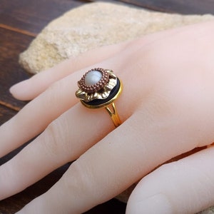 May include: A gold-colored ring with a central pearl-like stone, surrounded by a dark band and a decorative floral design. The ring is set on a hand, showcasing its intricate details and vintage style.