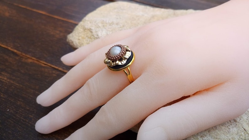 May include: A gold-colored ring with a central pearl-like stone, surrounded by a dark band and a decorative floral design. The ring is set on a hand, showcasing its intricate details and vintage style.