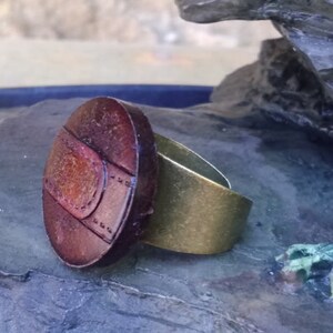 Op de afbeelding: Een ring met een grote, ovale, bruine hars steen met een gouden band. De steen heeft een getextureerd oppervlak met een patroon van lijnen en cirkels.