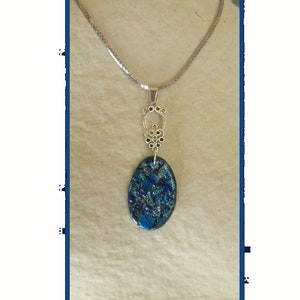 May include: A silver-toned necklace with an oval pendant. The pendant features a blue and black speckled design. The necklace has a decorative silver bail and a delicate chain.