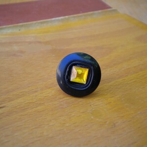 Puede incluir: Un anillo negro con una gema cuadrada amarilla en el centro. El anillo tiene forma circular con una incrustaci&oacute;n cuadrada. El anillo est&aacute; sobre una superficie de madera.