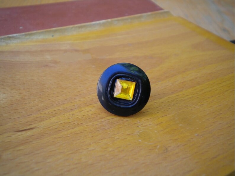 Puede incluir: Un anillo negro con una gema cuadrada amarilla en el centro. El anillo tiene forma circular con una incrustaci&oacute;n cuadrada. El anillo est&aacute; sobre una superficie de madera.