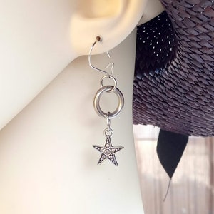 Può includere: Orecchini pendenti in argento con un ciondolo a forma di stella e un design a doppio anello. Gli orecchini sono appesi all'orecchio di una persona.