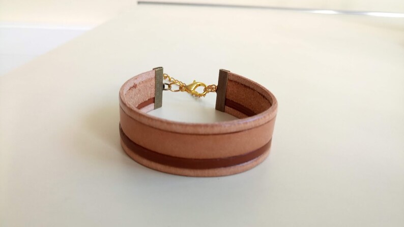 May include: A light brown leather bracelet with a brown stripe and a gold clasp.