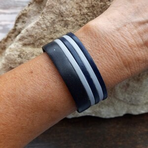 May include: A black leather bracelet with two white stripes. The bracelet is worn on a person's wrist.