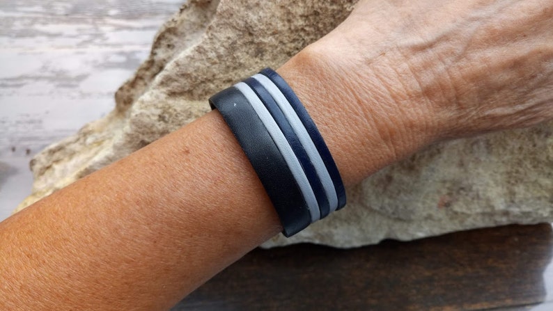 May include: A black leather bracelet with two white stripes. The bracelet is worn on a person's wrist.