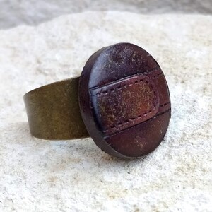 Op de afbeelding: Een bruine leren ring met een messing band. Het leer heeft een gestikt ontwerp en een verweerde look.
