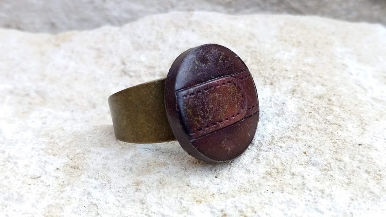 Op de afbeelding: Een bruine leren ring met een messing band. Het leer heeft een gestikt ontwerp en een verweerde look.