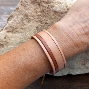 May include: A double-layered leather bracelet in shades of brown and beige. The bracelet is made of two strips of leather that are woven together.