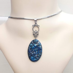 May include: A silver-toned necklace with a pendant on a mannequin. The pendant has an oval, blue and black design with a decorative silver-toned bail. The necklace chain is a thin, flat, silver-toned snake chain.