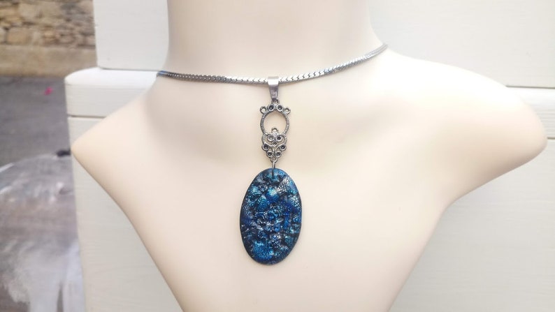 May include: A silver-toned necklace with a pendant on a mannequin. The pendant has an oval, blue and black design with a decorative silver-toned bail. The necklace chain is a thin, flat, silver-toned snake chain.