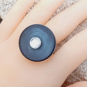 Puede incluir: Un anillo negro con una parte superior grande, redonda y plana. La parte superior del anillo tiene un pequeño strass transparente en el centro.