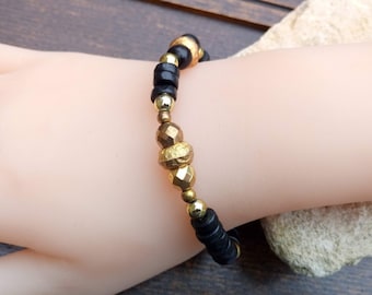 May include: A beaded bracelet featuring black and gold beads. The bracelet has a mix of round and faceted beads, with a central cluster of textured gold beads. The bracelet is displayed on a light-toned surface.