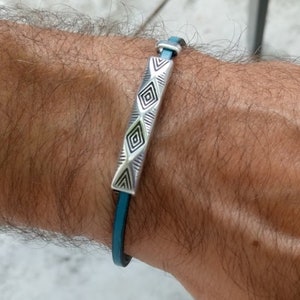 May include: A teal leather bracelet with a silver rectangular centerpiece. The centerpiece has geometric patterns and is connected to the leather band with silver hardware. The bracelet is designed to be worn on the wrist.