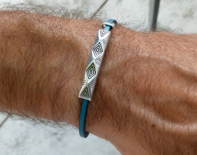 May include: A teal leather bracelet with a silver rectangular centerpiece. The centerpiece has geometric patterns and is connected to the leather band with silver hardware. The bracelet is designed to be worn on the wrist.