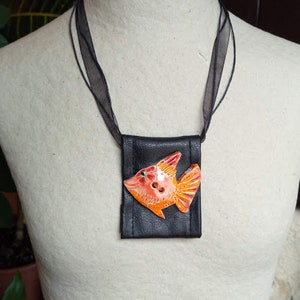 Pu&ograve; includere: Una collana in pelle nera con un piccolo sacchetto nero con un design di pesce arancione e rosso.