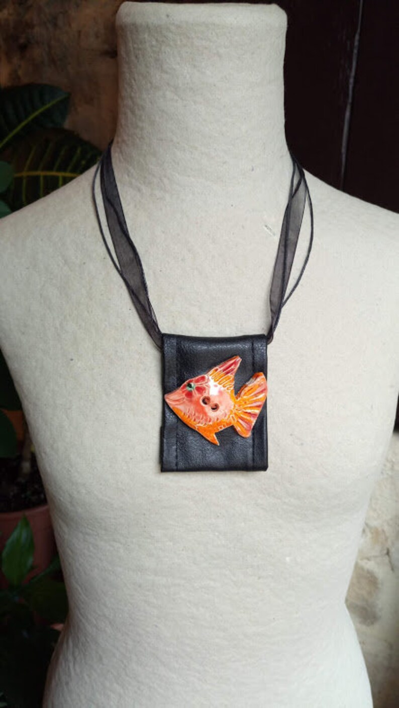 Pu&ograve; includere: Una collana in pelle nera con un piccolo sacchetto nero con un design di pesce arancione e rosso.