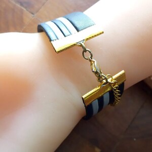 May include: A black and white striped leather bracelet with a gold clasp and chain. The bracelet is adjustable and has a simple design.