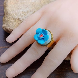 May include: A gold ring with a blue flower design. The flower has a pink center and is set in a clear resin.