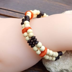 Può includere: Un braccialetto di perline a più fili. Il braccialetto presenta perline color crema, marrone scuro e arancione. Il braccialetto è indossato su un polso.