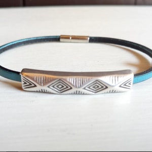 May include: A bracelet with a teal leather band and a black interior. It features a silver-coloured, rectangular centrepiece with a geometric pattern. The bracelet is secured with a silver-coloured clasp.