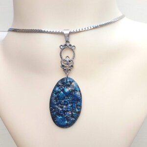 May include: A silver-toned necklace with a blue and silver oval pendant. The pendant has a textured, speckled appearance. The necklace is displayed on a mannequin.
