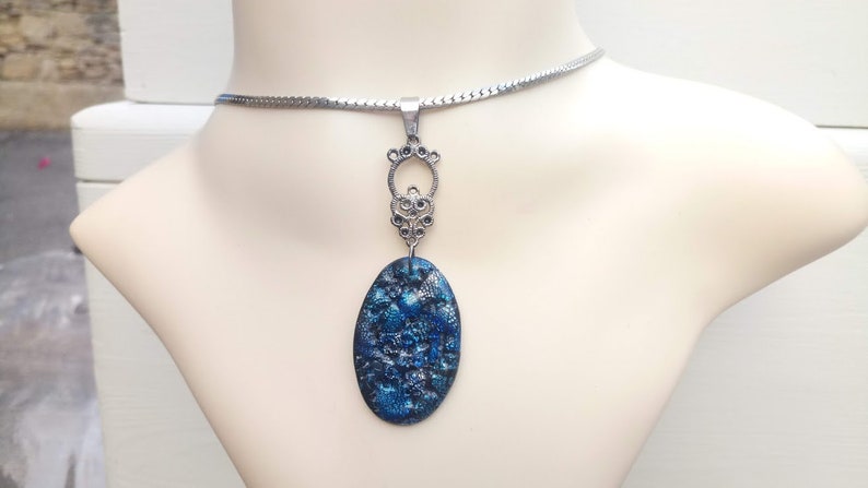 May include: A silver-toned necklace with a blue and silver oval pendant. The pendant has a textured, speckled appearance. The necklace is displayed on a mannequin.