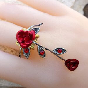 Può includere: Un anello in oro e argento con un design a forma di rosa rossa. L'anello ha una singola rosa rossa sulla fascia e una rosa rossa più piccola su uno stelo che si estende dalla fascia.