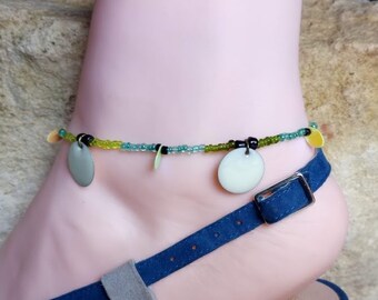 May include: A beaded ankle bracelet with green, blue and black beads and three silver-coloured charms. The bracelet is on a person's ankle.