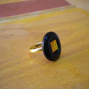 Puede incluir: Anillo dorado con un engaste negro de forma ovalada. El engaste presenta una piedra cuadrada amarilla y naranja. El anillo est&aacute; sobre una superficie de madera marr&oacute;n claro.