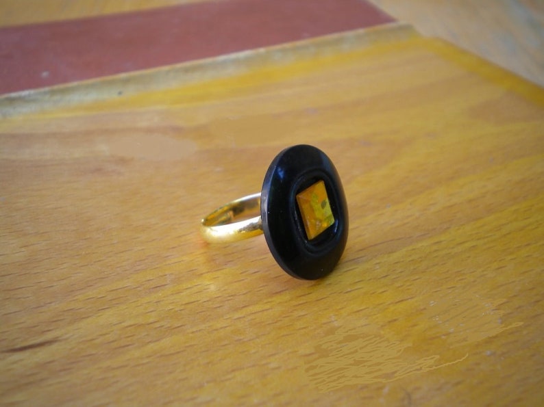 Puede incluir: Anillo dorado con un engaste negro de forma ovalada. El engaste presenta una piedra cuadrada amarilla y naranja. El anillo est&aacute; sobre una superficie de madera marr&oacute;n claro.