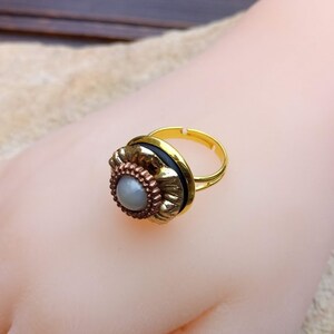 May include: A gold-toned adjustable ring with a floral design. The ring features a central gray stone surrounded by a copper-colored metal, a black band, and a gold-colored adjustable band. The ring is displayed on a mannequin hand.